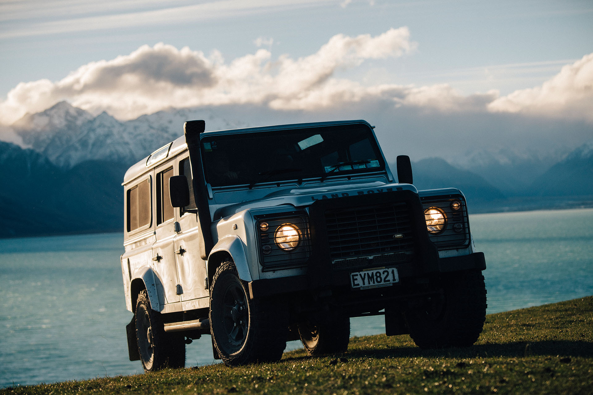 Land Rover with moody lighting, on a hillside with lake and snow capped mountains in the background